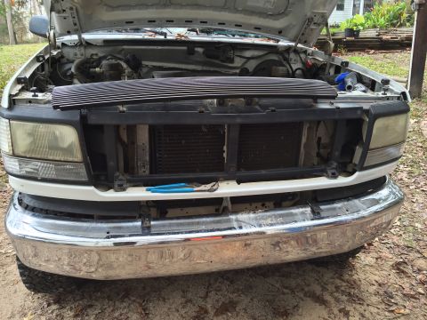 1996 Ford Bronco - The front end of a white 1996 Ford Bronco is shown with the hood open, revealing the engine bay. A new chrome grille is being installed ove…