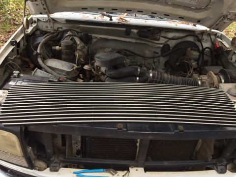 1996 Ford Bronco - The engine bay of a white 1996 Ford Bronco is shown with the hood open. A custom chrome grille is installed in front of the radiator.