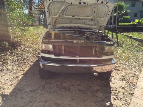 1996 Ford Bronco - The front of a white Ford Bronco with the hood open, revealing the engine bay. The vehicle is parked outdoors on a dirt and leaf-covered su…