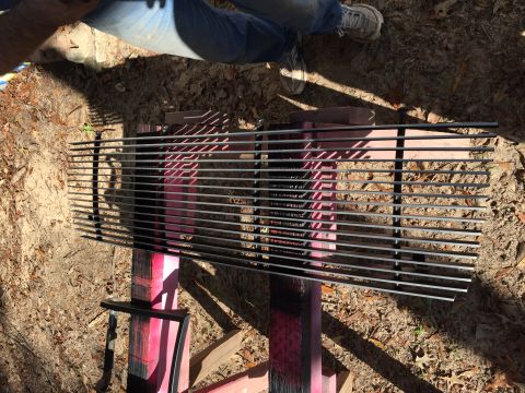 1996 Ford Bronco - A horizontal metal grille is laid out on pink and black spray-painted wooden supports outdoors. The grille is composed of many thin, parall…