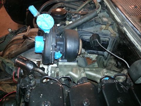 1994 Ford Bronco - A close-up view of a turbocharger installed in an engine bay, with blue tape covering some of the intake and exhaust ports. The engine comp…