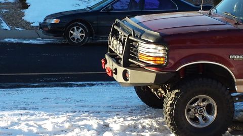 1996 Ford Bronco - A close-up, low-angle shot of the front of a dark red Ford Bronco with an aftermarket bumper and grille guard. The vehicle is parked on a s…
