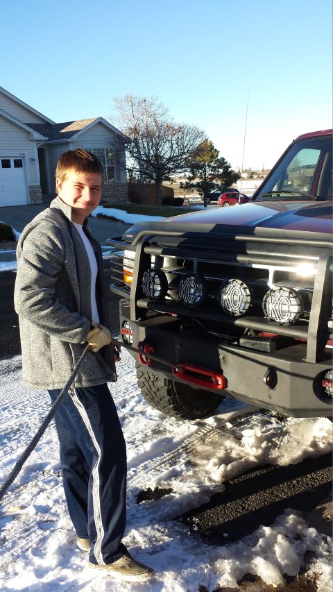 1996 Ford Bronco - A young man in a gray jacket and blue sweatpants stands next to the front of a large, dark red SUV, holding a tow rope. The SUV has a heavy…