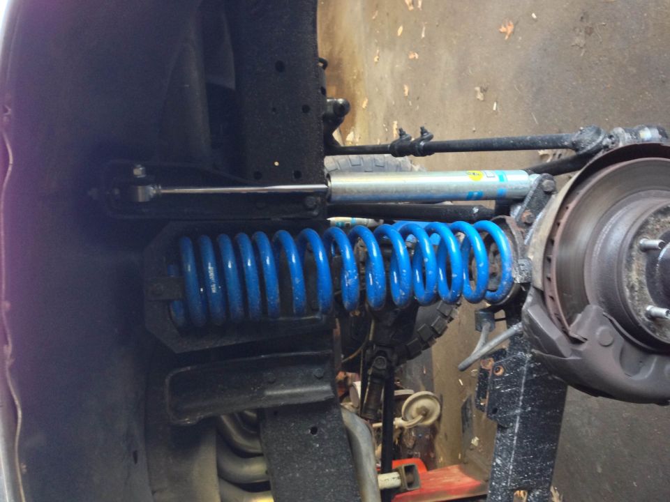 Close-up of blue coil spring and silver Bilstein shock on vehicle suspension with brake rotor.