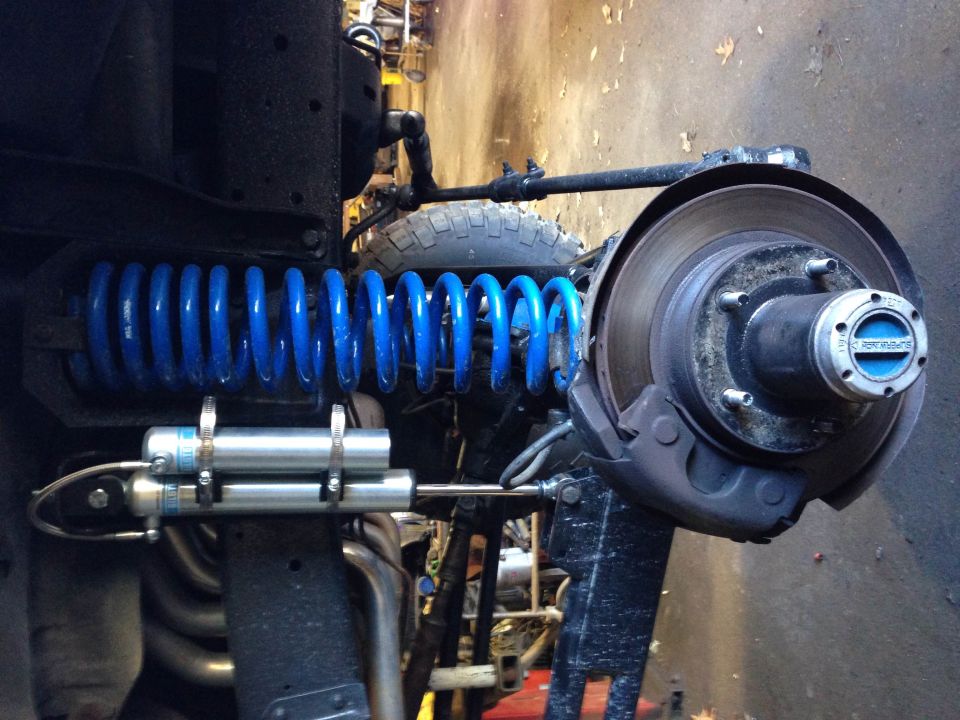 Close-up of vehicle front suspension with blue coil spring and Bilstein steering stabilizer.