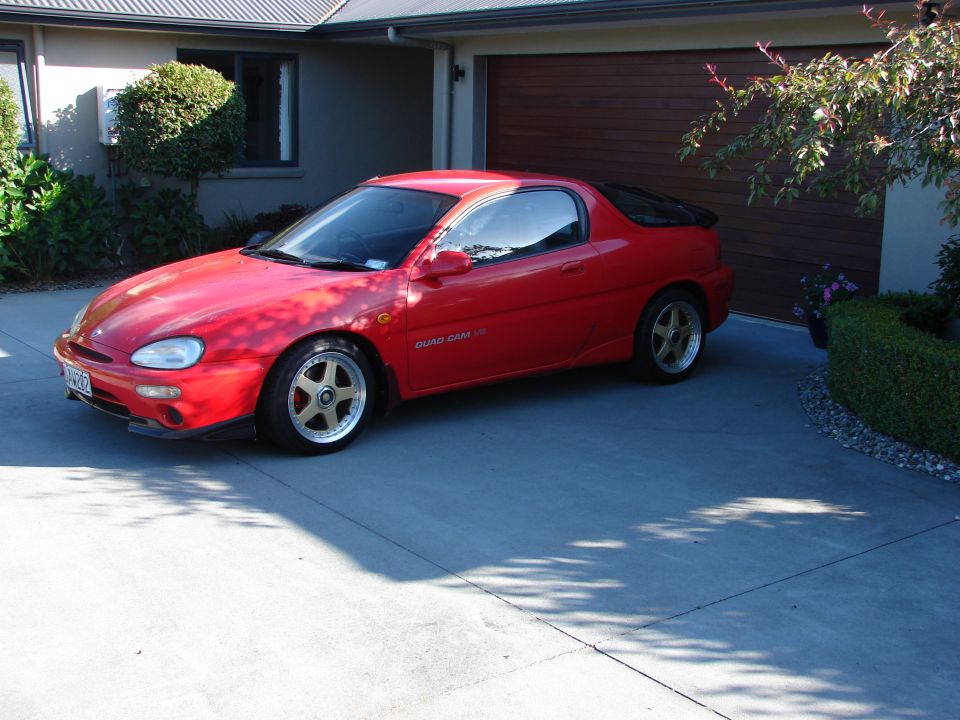 Red 1993 Mazda MX-3 coupe with gold wheels and black spoiler parked in front of a house.