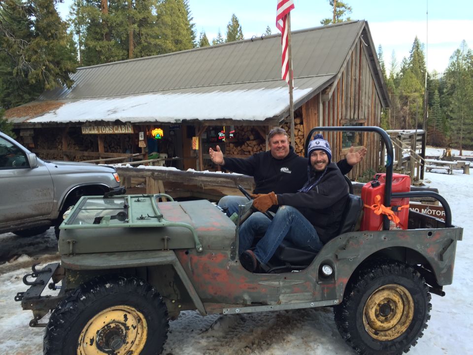 Old Jeep Willys MB with people in it, parked outside a building with snow on the roof. Another vehicle is to the left