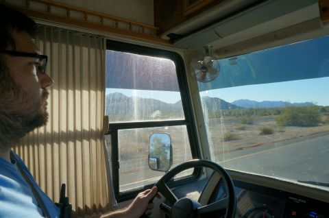 1995 Chevrolet P30 1 Ton - Step Van - A man wearing glasses drives a motorhome, looking out the windshield at a desert landscape with mountains in the distance. The interior sho…