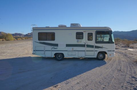 1995 Chevrolet P30 1 Ton - Step Van - Side profile of a white Fleetwood Flair Class A motorhome with green graphics parked on desert ground with mountains behind.