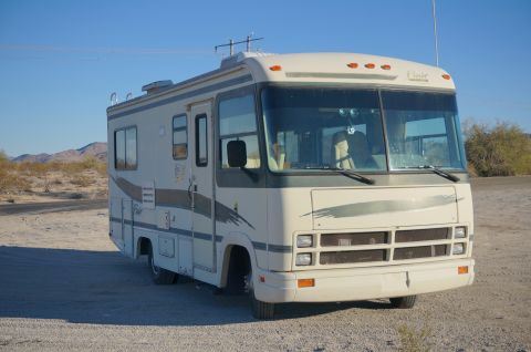1995 Chevrolet P30 1 Ton - Step Van - A beige and gray Fleetwood Flair RV is parked on a dirt lot with desert scrub and mountains in the background under a clear blue sky.