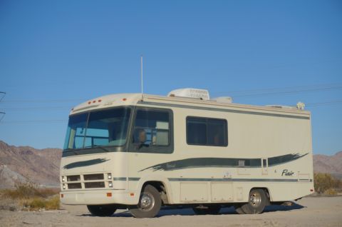 1995 Chevrolet P30 1 Ton - Step Van - A cream-colored RV with dark green graphics is parked on a dirt lot with mountains and a clear blue sky in the background. The RV has multi…
