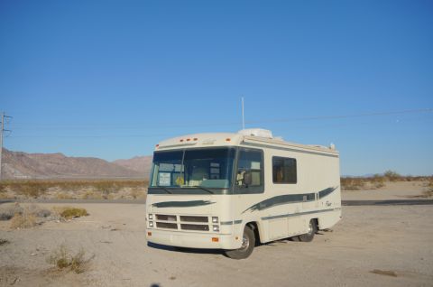 1995 Chevrolet P30 1 Ton - Step Van - A cream-colored Class A RV with dark green pinstriping is parked in a dirt lot under a clear blue sky. The RV has a large windshield, side…