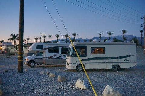 1995 Chevrolet P30 1 Ton - Step Van - Two recreational vehicles are parked on a gravel lot with palm trees and mountains in the background during twilight. The RV in the foregro…