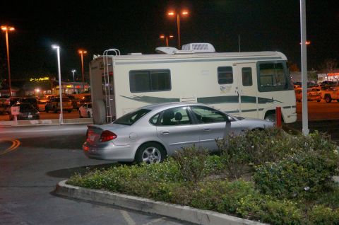 1995 Chevrolet P30 1 Ton - Step Van - A silver sedan is parked in front of a large RV at night. The RV is parked in a lot with streetlights and other vehicles visible in the bac…