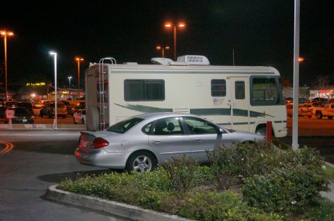 1995 Chevrolet P30 1 Ton - Step Van - A silver sedan is parked in front of a beige RV at night. The RV has a ladder attached to the back and is parked in a lot with streetlights.