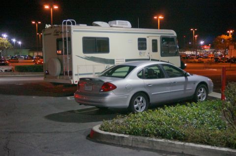1995 Chevrolet P30 1 Ton - Step Van - A silver sedan is parked next to a large RV at night in a parking lot. The RV is white with beige accents and has a spare tire mounted on t…