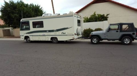 1995 Chevrolet P30 1 Ton - Step Van - A beige Fleetwood Flair RV is towed by a gray Jeep Wrangler. The RV is parked on a paved street in front of a residential building.