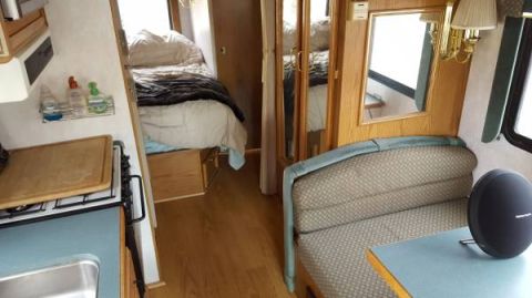 1995 Chevrolet P30 1 Ton - Step Van - The interior of a vehicle shows a bed with dark bedding and white linens, a small kitchen area with a cutting board on a stovetop, and a se…