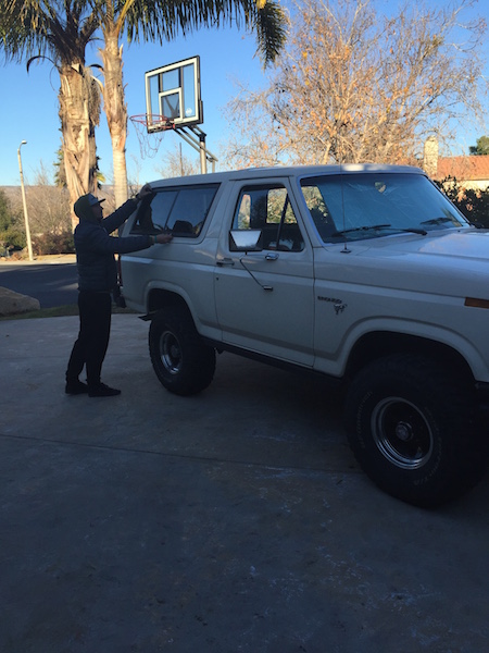 1981 Ford Bronco - A person in a dark jacket and hat is reaching towards the roof of a white Ford Bronco parked on a concrete driveway. A basketball hoop is v…