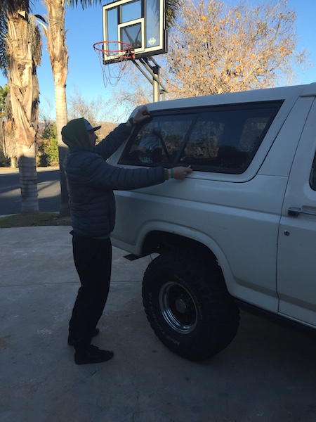 1981 Ford Bronco - A person in a gray puffer jacket and black pants is adjusting the window of a white Ford Bronco. A basketball hoop is visible in the backgr…