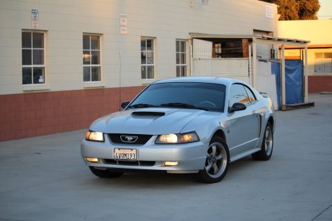 2002 Ford Mustang - A silver 2002 Ford Mustang is parked in front of a building with a brick base.