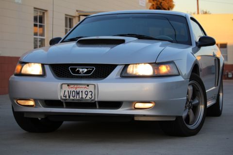 2002 Ford Mustang - A low-angle, front-facing shot of a silver Ford Mustang with its headlights on. The California license plate reads "4VQM193".