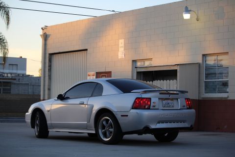 2002 Ford Mustang - A silver 2002 Ford Mustang GT coupe is parked in front of a brick building with a large garage door. The rear of the car is visible, showin…