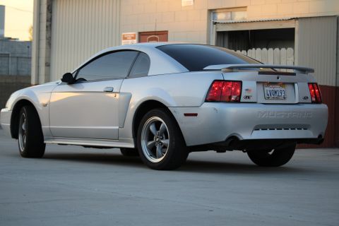 2002 Ford Mustang - The rear passenger side quarter view of a silver Ford Mustang GT coupe is shown parked on a concrete surface in front of a building. The ca…