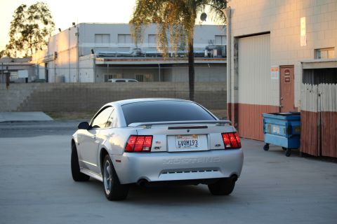 2002 Ford Mustang - Rear 3/4 view of a silver Ford Mustang coupe parked on a concrete surface in front of a building with a blue dumpster nearby.