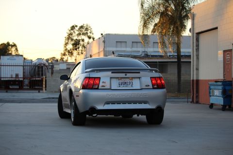 2002 Ford Mustang - The rear view of a silver Ford Mustang is shown parked on a concrete surface in front of a building with a large garage door. The car has a…