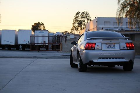 2002 Ford Mustang - The rear view of a silver Ford Mustang GT is parked in a lot with trailers and buildings in the background during sunset.