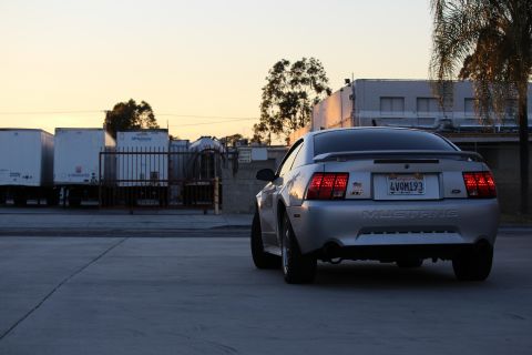 2002 Ford Mustang - The rear view of a silver Ford Mustang GT is shown parked in an industrial area at sunset. The car has a spoiler and a California license p…