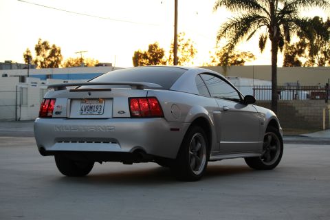 2002 Ford Mustang - The rear driver's side quarter view of a silver Ford Mustang coupe is shown parked on a concrete surface. The car has a spoiler, a Californ…