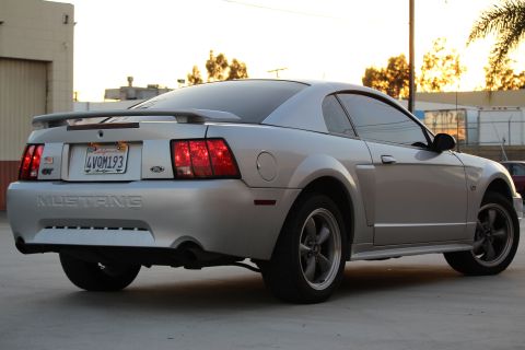 2002 Ford Mustang - The rear driver's side quarter view of a silver Ford Mustang GT coupe is shown parked on concrete with a building and trees in the backgrou…