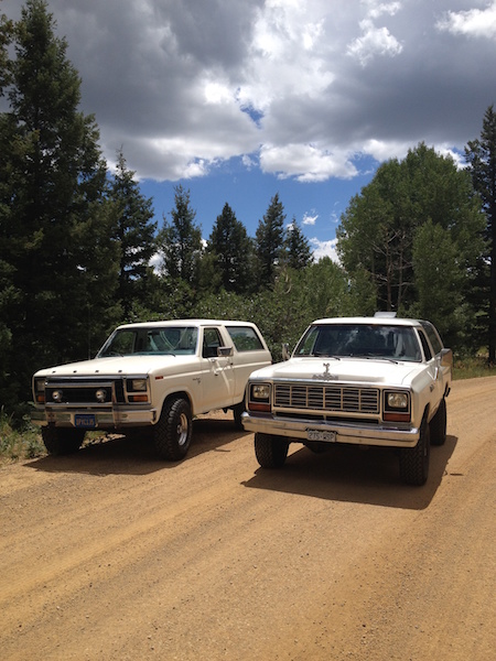 1981 Ford Bronco - Two white SUVs are parked on a dirt road in a wooded area. The vehicle on the left is a Ford Bronco and the vehicle on the right appears to…