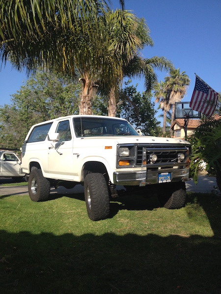 1981 Ford Bronco - A lifted white 1980s Ford Bronco is parked on a grassy lawn in front of palm trees and a house. The vehicle has large off-road tires and ch…