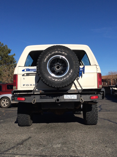 1981 Ford Bronco - The rear view of a white 1980s Ford Bronco is shown parked outdoors under a clear blue sky. It features a spare tire mounted on the back, a…