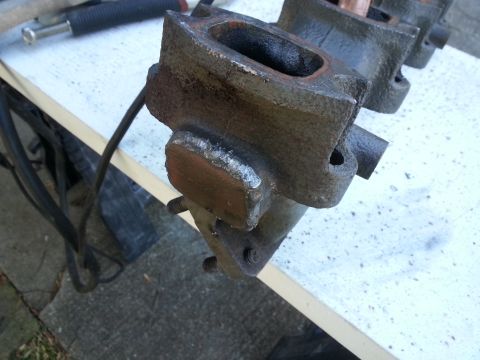 1994 Ford Bronco - A close-up view of a welded metal part, likely an exhaust manifold, resting on a white surface. The part is dark gray with some rust and sh…