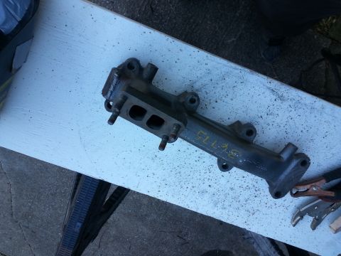 1994 Ford Bronco - A dark gray exhaust manifold with yellow markings is shown lying on a white surface. The manifold has two visible ports and several bolt st…