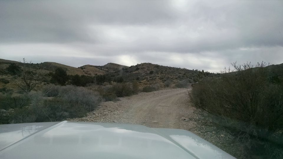 White vehicle hood and windshield looking at a dirt road in a hilly, arid landscape.