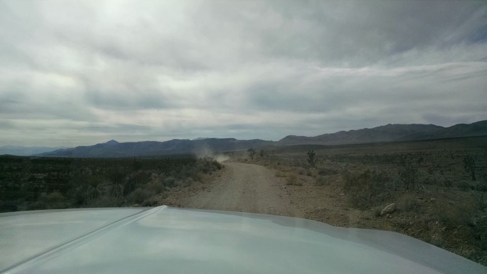 White vehicle hood in foreground, looking at a dusty desert road and mountains.