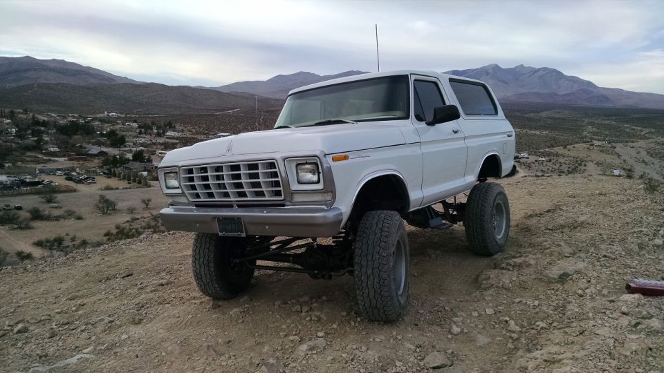 Lifted white 1979 Ford Bronco parked on a dirt hill with a town and mountains in the background.
