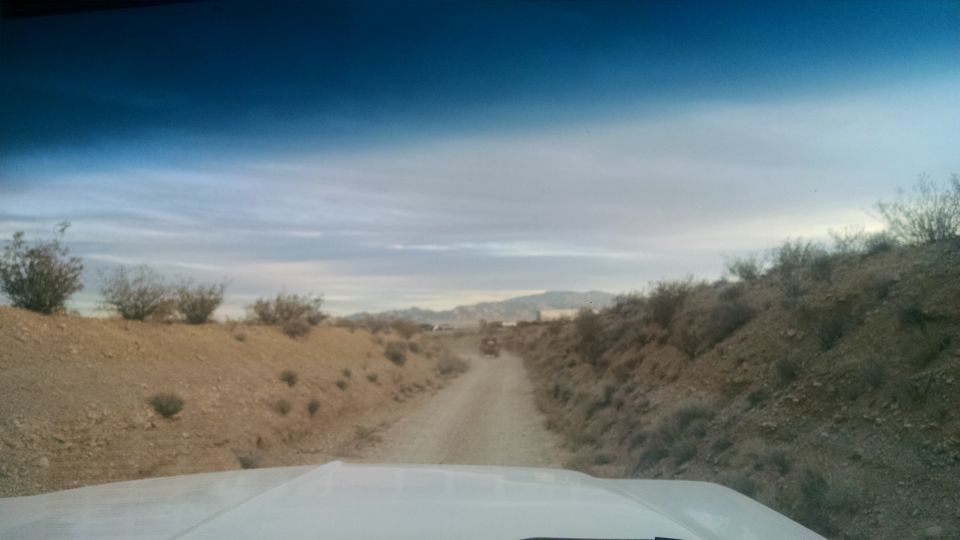 View from inside a white vehicle looking down a dirt road in a desert with two off-road vehicles ahead.