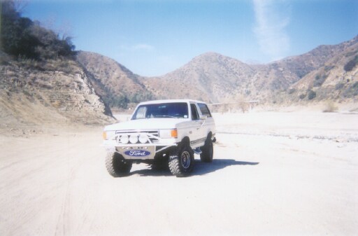 White lifted Ford Bronco SUV parked in a dry sandy riverbed with mountains behind.