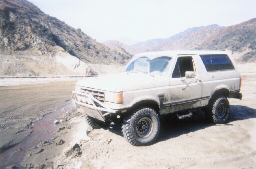 White two-door Ford Bronco SUV on dirt riverbed with mountains behind