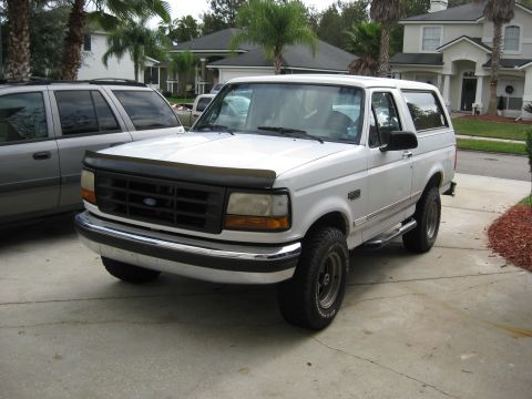1994 Ford Bronco - A white 1994 Ford Bronco is parked on a concrete driveway in front of a house. The SUV is viewed from a 3/4 front angle, showing its chrome…