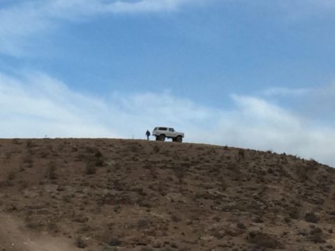 1979 Ford Bronco - A lone SUV is seen driving along the crest of a barren, dusty hill under a partly cloudy sky.