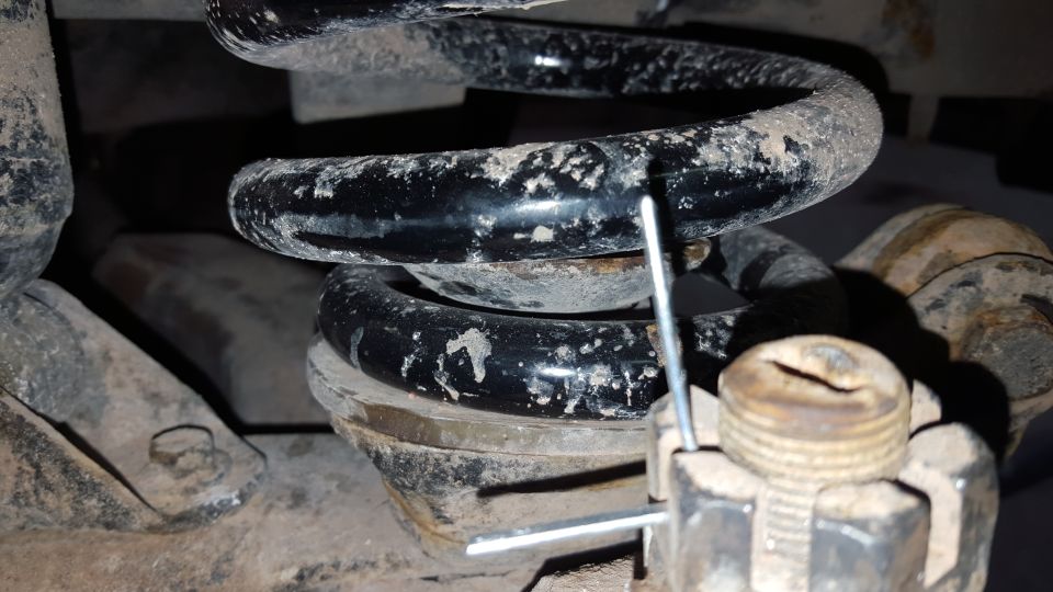 Close-up of a dirty black coil spring with a metal pin inserted, resting on a metal mount with a large nut