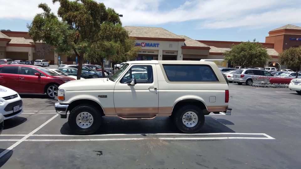 White and tan Ford Bronco SUV, side view, parked in an H Mart shopping center lot