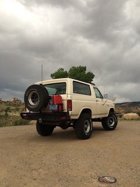 1981 Ford Bronco - This lifted Ford Bronco sits parked on a dirt lot under a cloudy sky, equipped with a rear-mounted spare tire and red fuel cans.